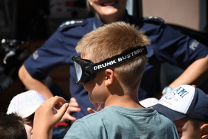 W tle stojąca, uśmiechnięta policjantka a przed nią dziecko z założonymi alkogogle.